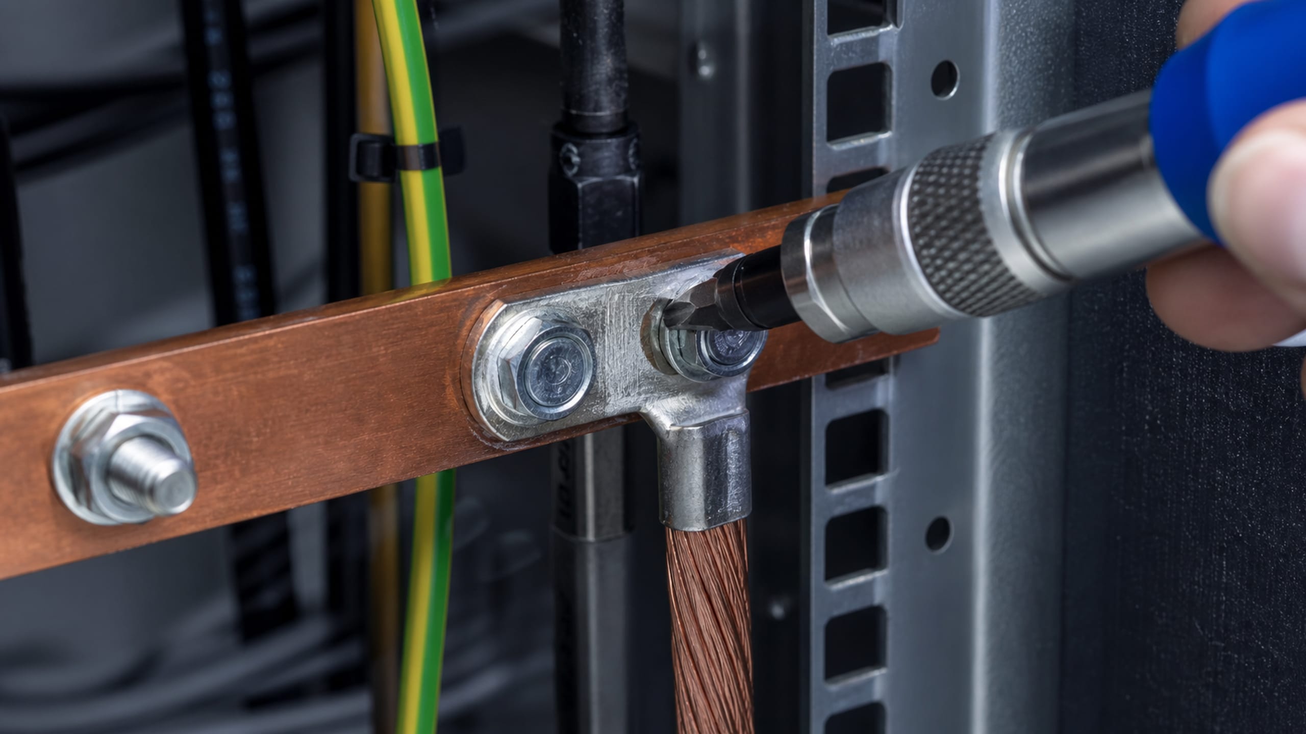 A torque driver tightening a braided bonding conductor onto a copper telecommunications grounding busbar inside a rack.